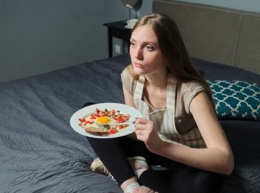 Girl sitting on bed with remote control watching TV and eating síntomas de los trastornos alimentarios